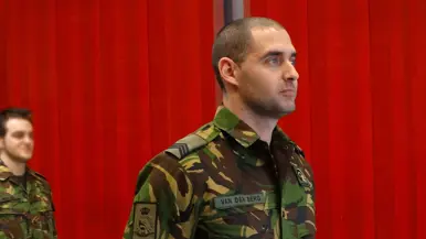 Sergeant 1 Erik staat in de houding voor een rode schuifwand, met in de achtergrond een collega.