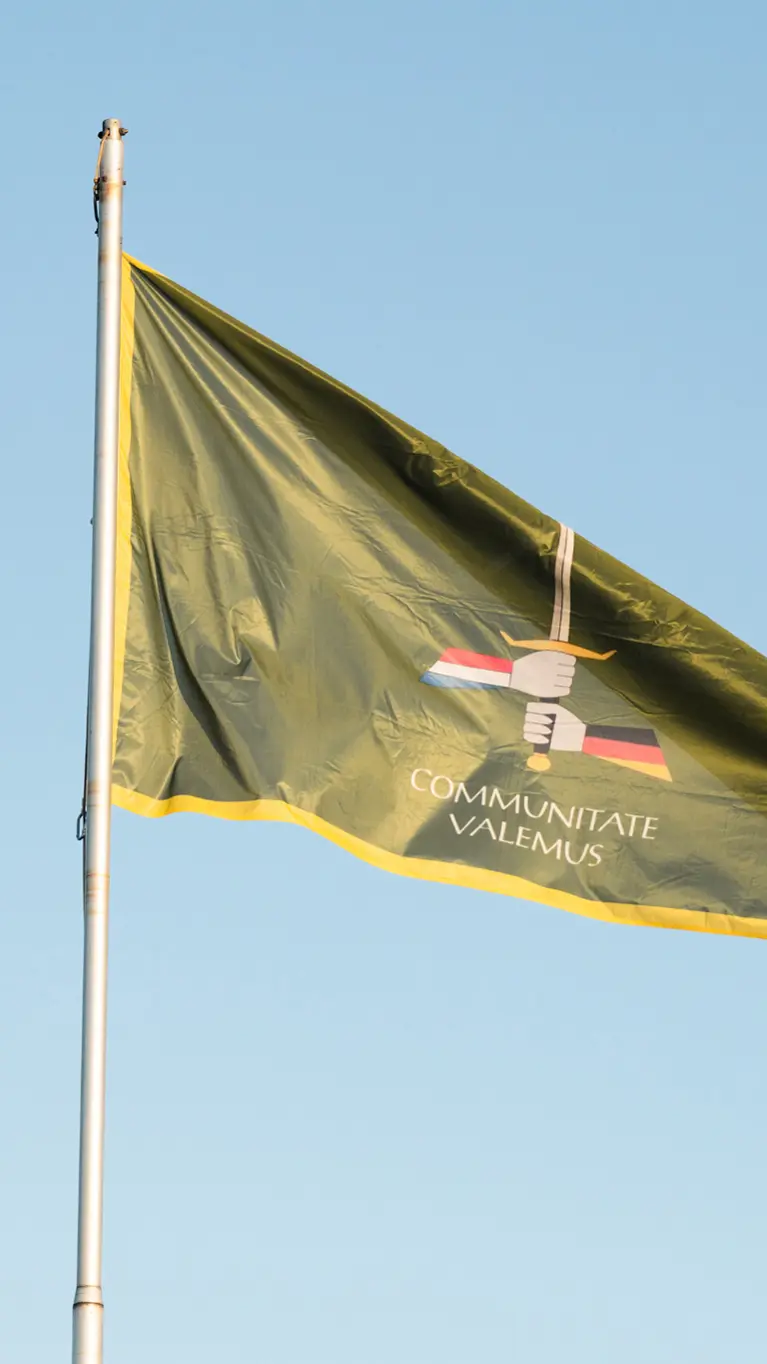 Wapperende, groene vlag met daarop het embleem van 1GNC. Groen wapenschild met daarop een zwaard dat wordt vastgehouden door een 'Duitse' en een 'Nederlandse' hand. De spreuk ‘Communitate valemus’ betekent ‘samen zijn we sterk’. 