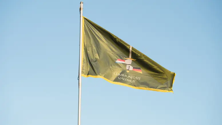 Wapperende, groene vlag met daarop het embleem van 1GNC. Groen wapenschild met daarop een zwaard dat wordt vastgehouden door een 'Duitse' en een 'Nederlandse' hand. De spreuk ‘Communitate valemus’ betekent ‘samen zijn we sterk’. 