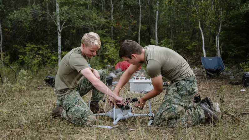 2 militairen in t-shirt buigen zich op grasveld in bos over een drone.