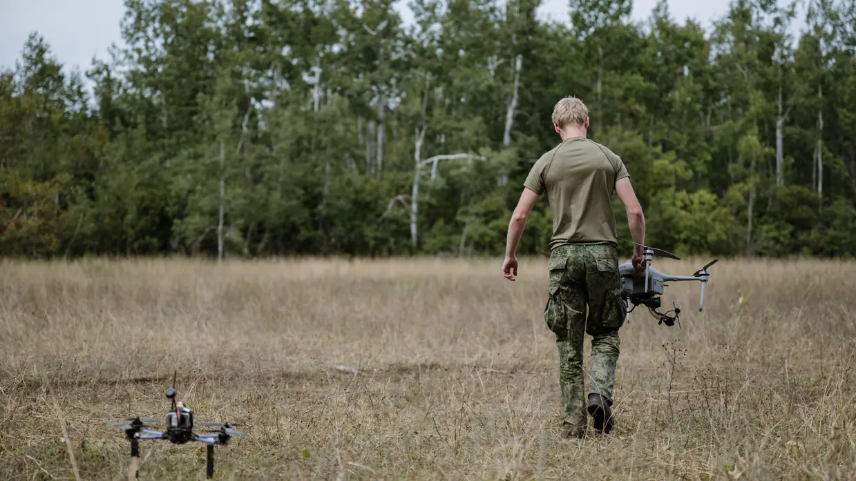 Een drone staat op grasveld in bos terwijl een militair wegloopt met een onbemenste mini-drone in handen.