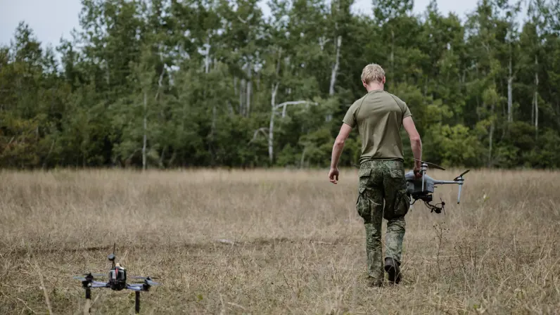 Een drone staat op grasveld in bos terwijl een militair wegloopt met een onbemenste mini-drone in handen.