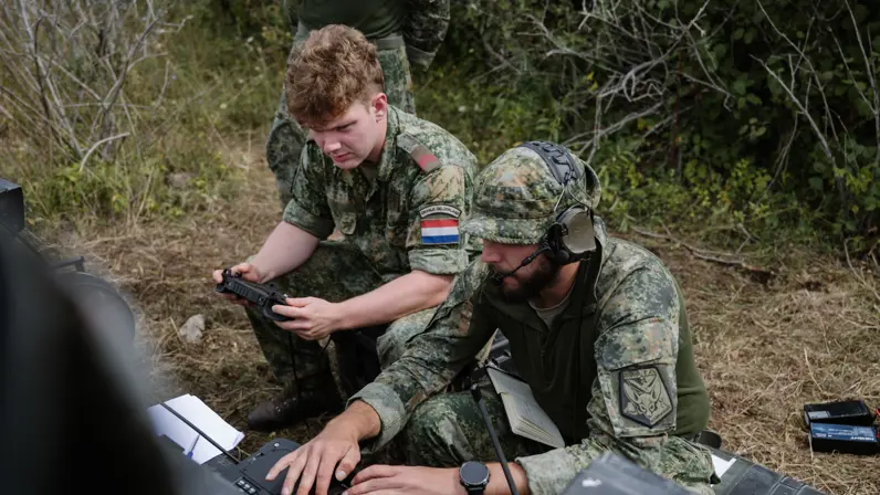 2 militairen kijken zittend in een bos op FPV-monitor en een beeldscherm.