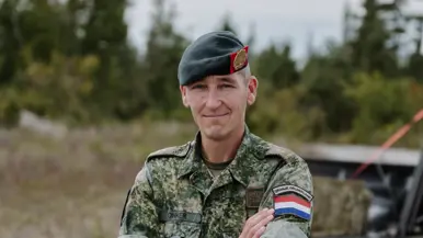 Een militair met baret op poseert met armen over elkaar met achter hem 2 zittende militairen.