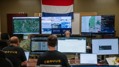 3 heren houden in een soort studio allerlei beeldschermen in de gaten met op achtergrond de Nederlandse vlag.