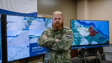 Een militair met baard poseert met armen over elkaar voor 2 grote beeldschermen en een deel van de Nederlandse vlag.