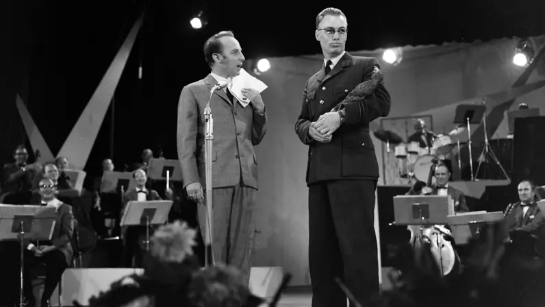  Een man spreekt op het toneel in een microfoon met papier in hand, naast hem een militair met een trofee in zijn arm. Achter hen speelt een orkest onder spotlights; het publiek kijkt als vage silhouetten.