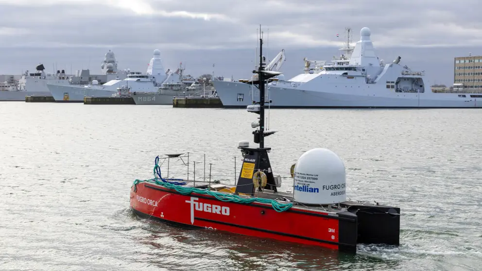 Een varend rood vaartuig dat op afstand bestuurbaar is met marineschepen op de achtergrond.