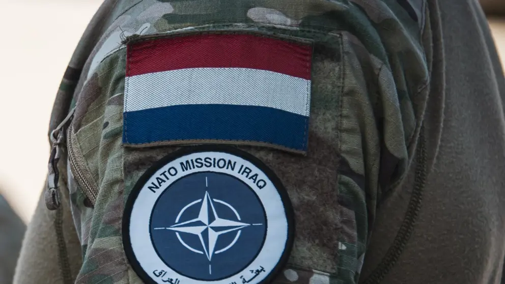 Schouder militair met NAVO-embleem en Nederlandse vlag erboven.