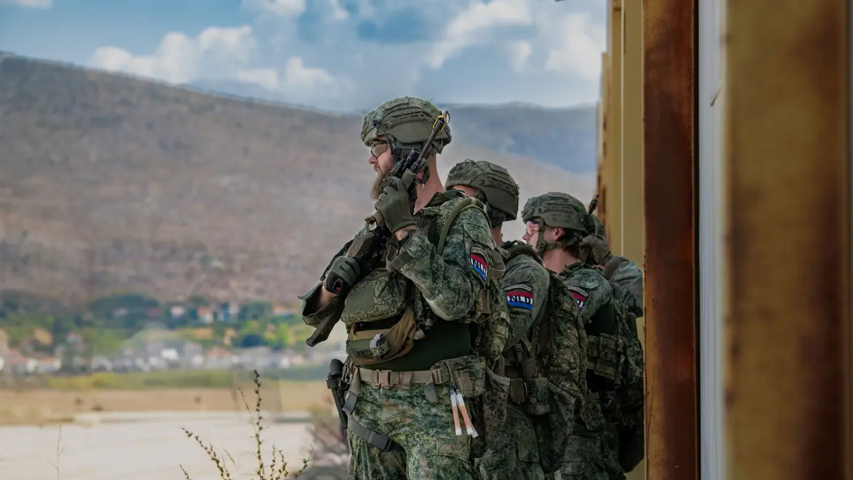 Militairen met wapen in de hand staan achter elkaar langs hoog hek.