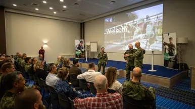 Zaal met zittende militairen van verschillende krijgsmachtdelen en enkele burgers. Ze kijken naar een podium met een projectiescherm. Daarvoor staat generaal Onno Eichelsheim samen met twee collega-militairen.