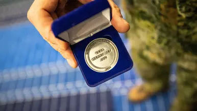 Een zilverkleurige munt met de tekst ‘Instructeursdag DCPL 2026; VeVa werkt met jou’. Deze wordt door een hand van een militair getoond in een blauw doosje.