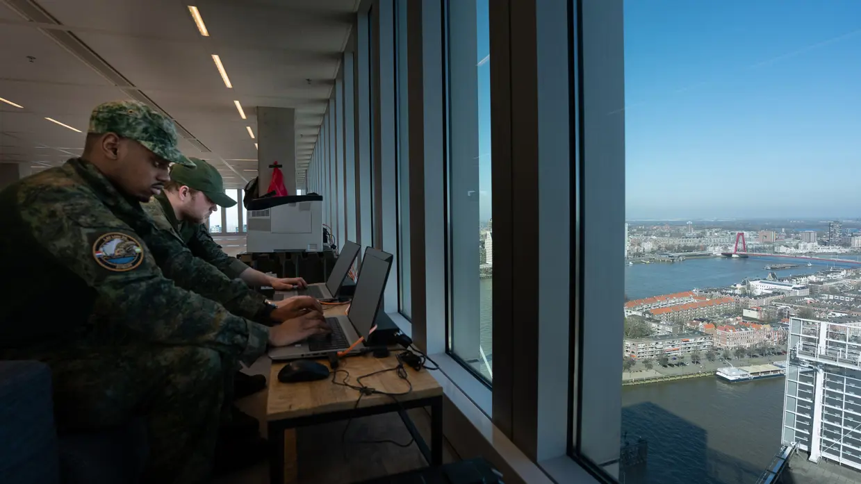 Twee militairen werken achter hun laptop. Het duo kijkt vanaf hun positie op hoogte uit op Rotterdam.