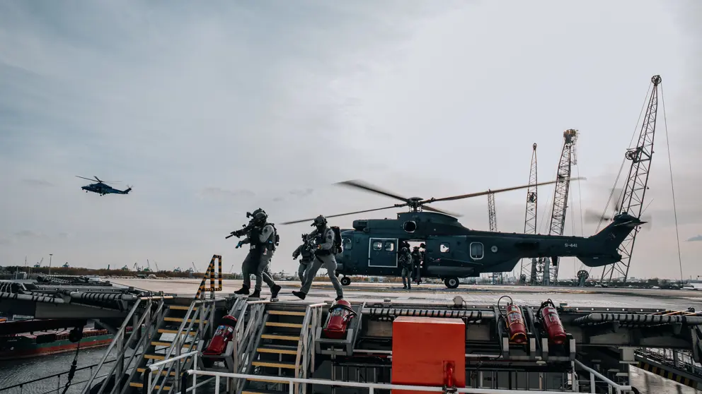 Apache-gevechtshelikopter op platform. Militairen met getrokken wapens ervoor. Verder weg een Apache in de lucht.