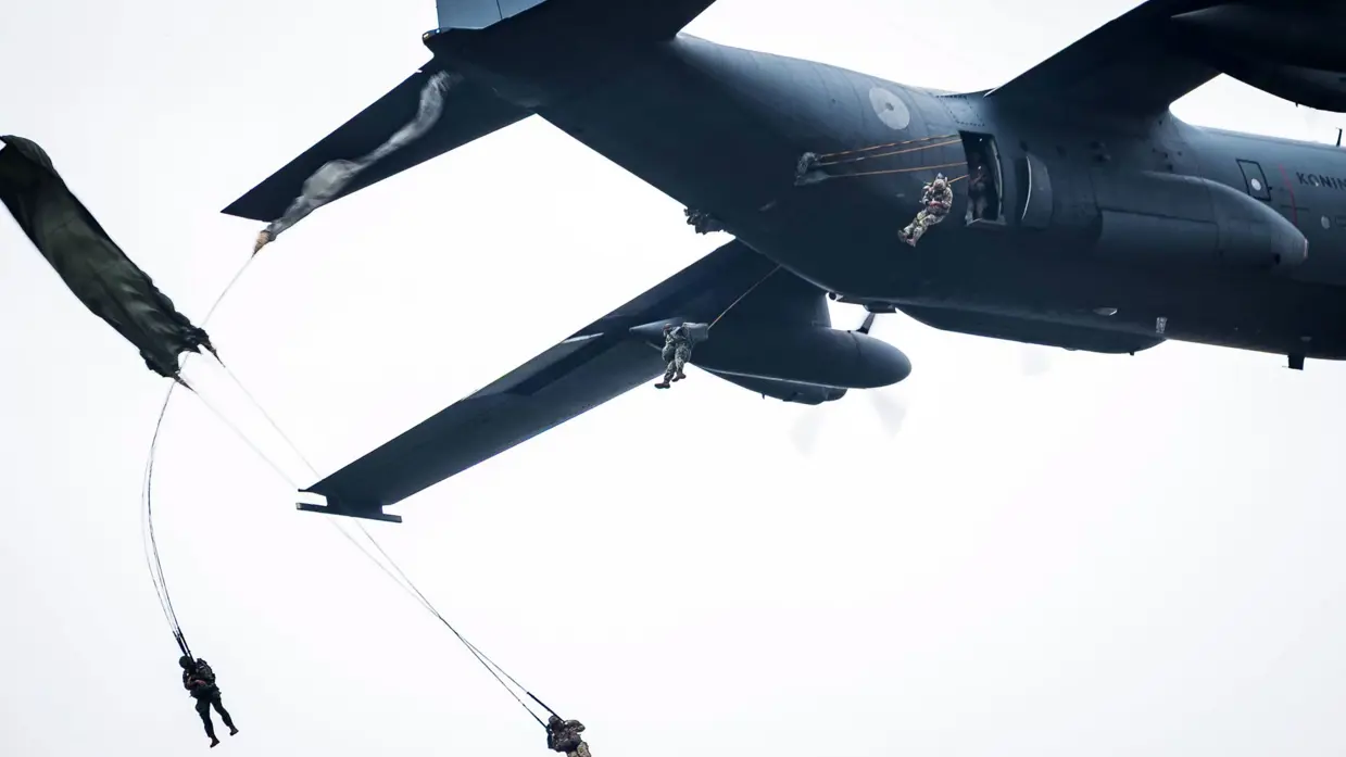 Een groot militair vliegtuig met staartnummer G-781 vliegt door de bewolkte lucht. De achterlijke laadklep van het vliegtuig staat open, en daarbovenuit springen meerdere soldaten in camouillagekleding aan parachutes naar beneden.