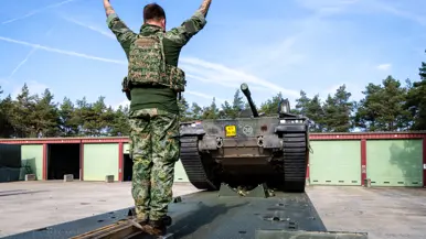 Een soldaat in camouflageuniform staat op een aanhangwagen, armen geheven. Voor hem een gepantserd rupsvoertuig met geschutstorens. Blauwe lucht, rode en groene gebouwen op achtergrond.
