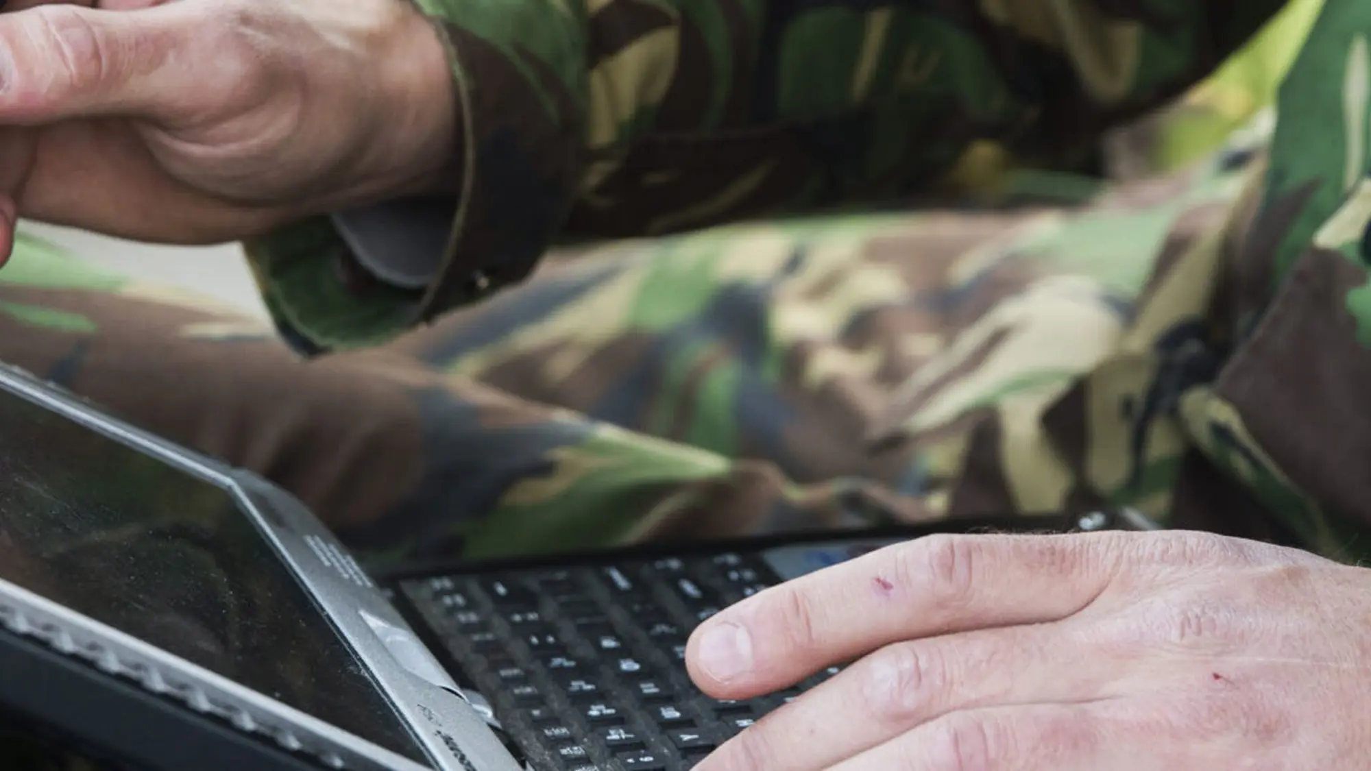 Close-up van 2 handen van militair die tablet op schoot heeft. 1 hand wijst met een pen op de tablet, de andere hand typt.
