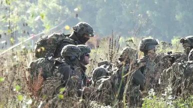 Soldaten in camouflage met helmen en rugzakken zitten gehurkt in hoog, droog gras. Achter hen een groene helling.