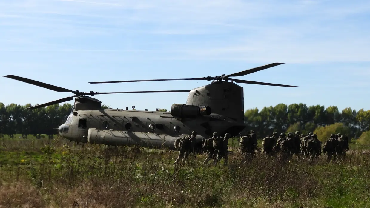 Een grote groene Chinook-helikopter met dubbele rotoren staat in hooggras. Soldaten in camouflage lopen in een lijn ernaartoe. Achter hen bomen; helderblauwe lucht.