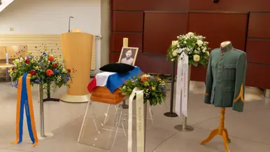 : De grafkist staat opgesteld in een ruimte met kransen, een foto en een oud uniform.