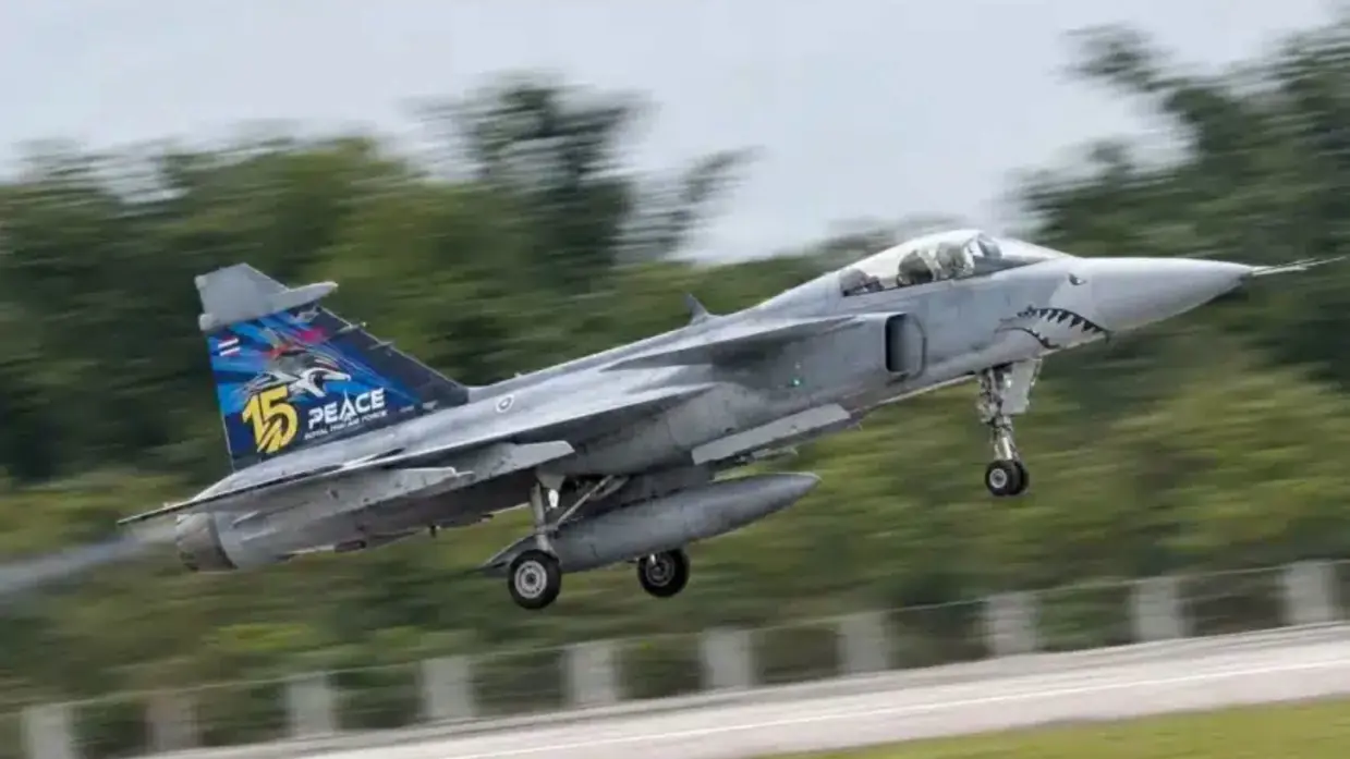 Een grijs gevechtsvliegtuig (Gripen) vliegt laag over de landingsbaan met uitgeklapt landingsgestel. De staart toont nummer "15" met "PEACE" als opschrift. Groene bomen, grijze lucht.