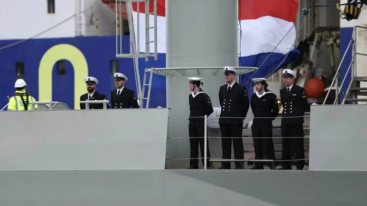 Bemanning staat op het dek. Op de achtergrond de Nederlandse driekleur.