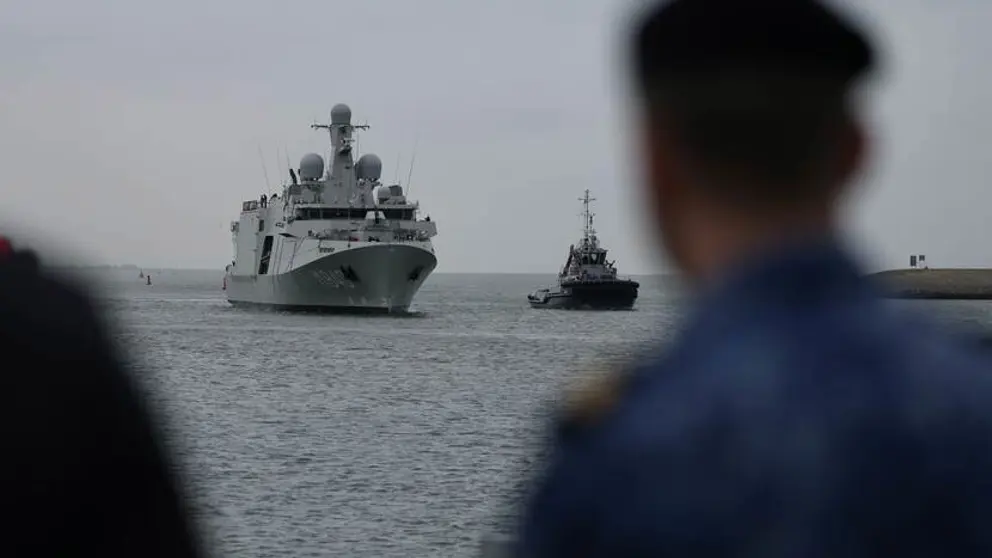 Het grijze schip De Vlissingen komt aangevaren. Rechts vaag een marinier in beeld.