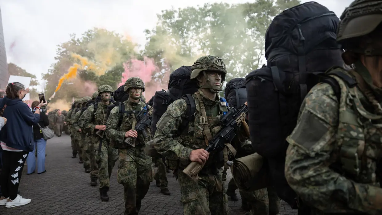 Militairen met grote rugtassen, een geweer voor de borst en camouflagehelm op marcheren over straat langs burgers met handfakkels met gekleurde rook.