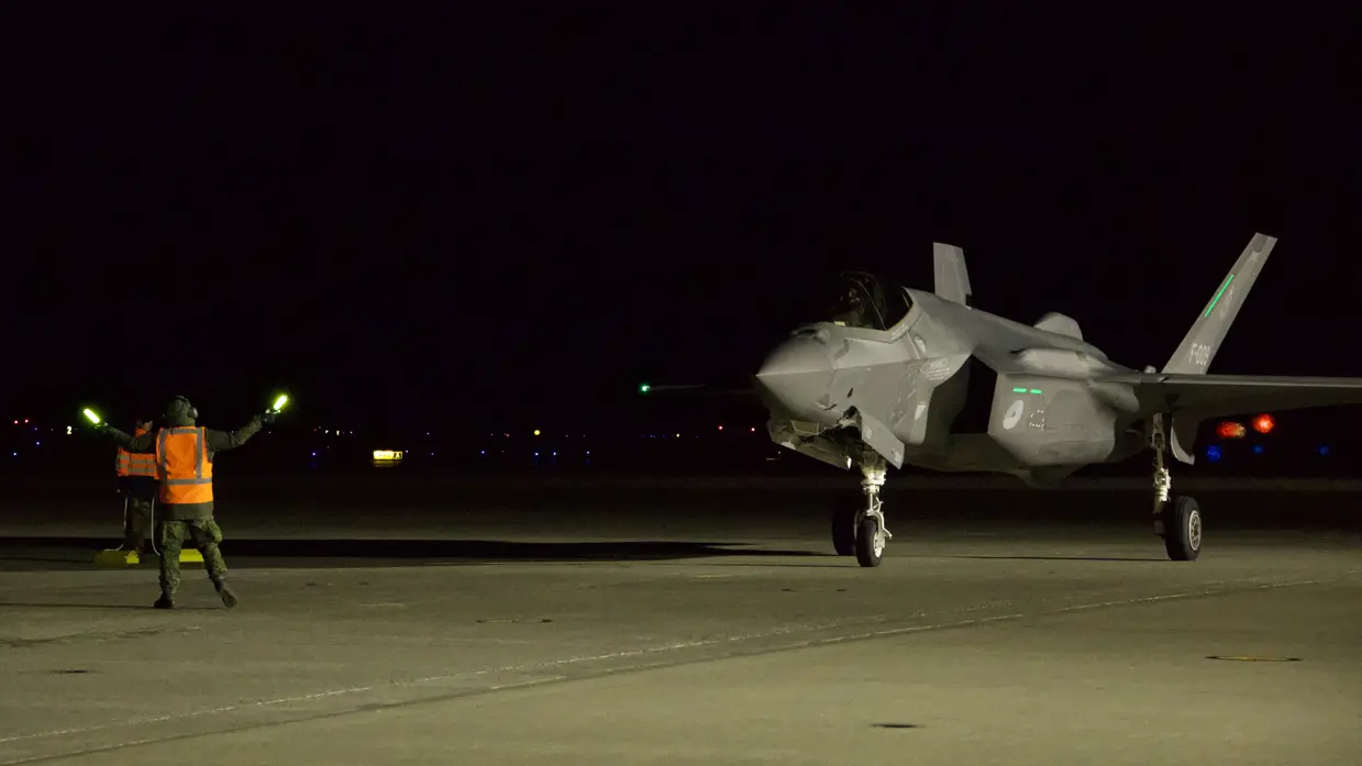 In het donker loodst grondpersoneel met een oranje hesje aan en met lichtgevende sticks in de handen een F-35 naar zijn parkeerplaats.