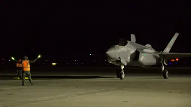In het donker loodst grondpersoneel met een oranje hesje aan en met lichtgevende sticks in de handen een F-35 naar zijn parkeerplaats.