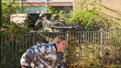 Vooraan loopt iemand in camouflagejas met een appel; erachter een tactisch voertuig met twee gehelmde soldaten. Stenen gebouw met hek en groen eromheen.