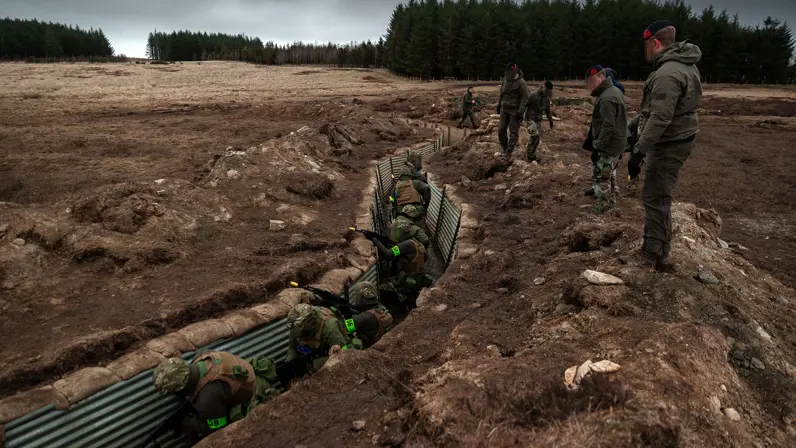 Alt-tekst: Militairen verplaatsen zich achter elkaar door een loopgraaf. Van bovenaf worden zij geobserveerd door instructeurs. 
