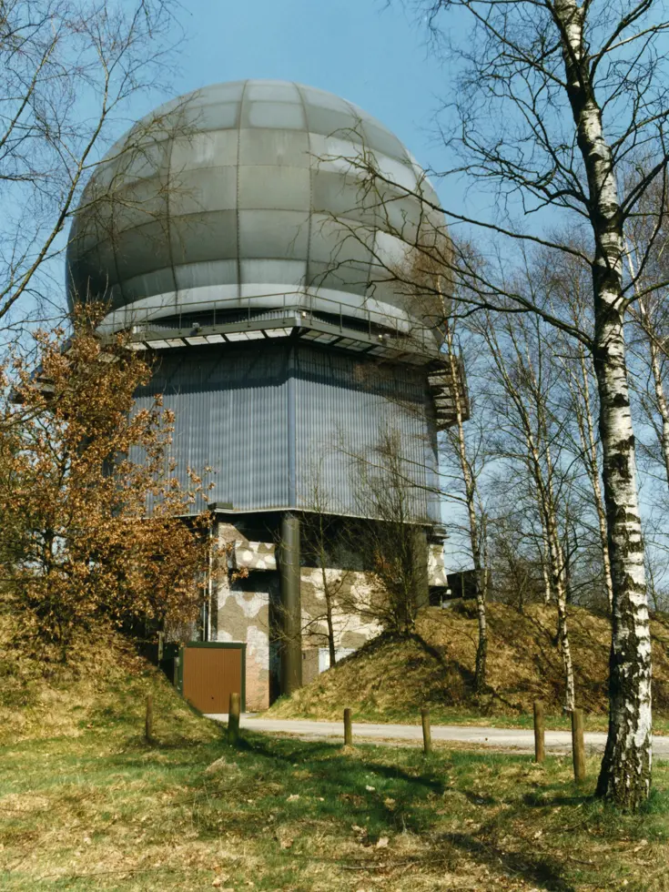 Een radarbol op een laag gebouw met links begroeiing en rechts een boom.
