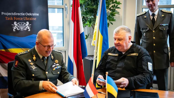 Kolonel Simon Wouda en chief engineer Vadym Nurmahomedov tekenen zittend aan een tafel de overdracht. Achter hen de Nederlandse en Oekraïense vlag.