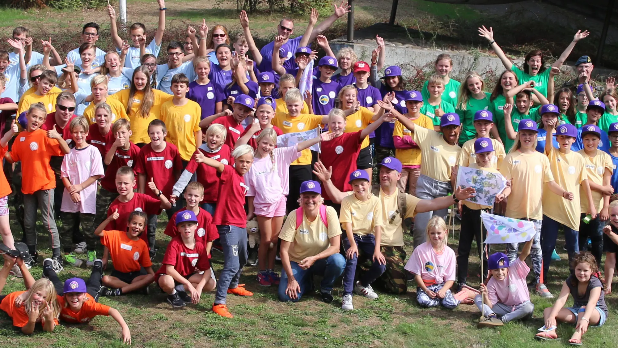 Kinderen, die allemaal verschillende kleuren dragen, zwaaien enthousiast naar de camera.