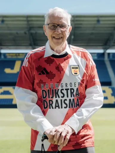 Een glimlachende oudere man staat op een voetbalveld, gekleed in een rood-wit shirt met esdoornbladeren en de tekst "Dijkstra Draisma" en "Bouwgroep" erop.