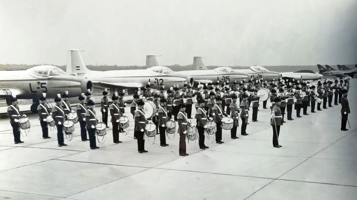 Het orkest staat opgesteld op een taxibaan, met in de achtergrond diverse luchtmachtvliegtuigen.