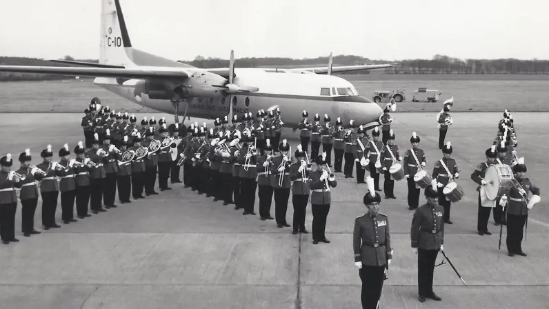 Foto op een vliegveld: een militaire kapel in uniform met hoge hoeden en witte pluimen staat in formatie met trompetten, trombones en drums; op de achtergrond staat een transportvliegtuig.