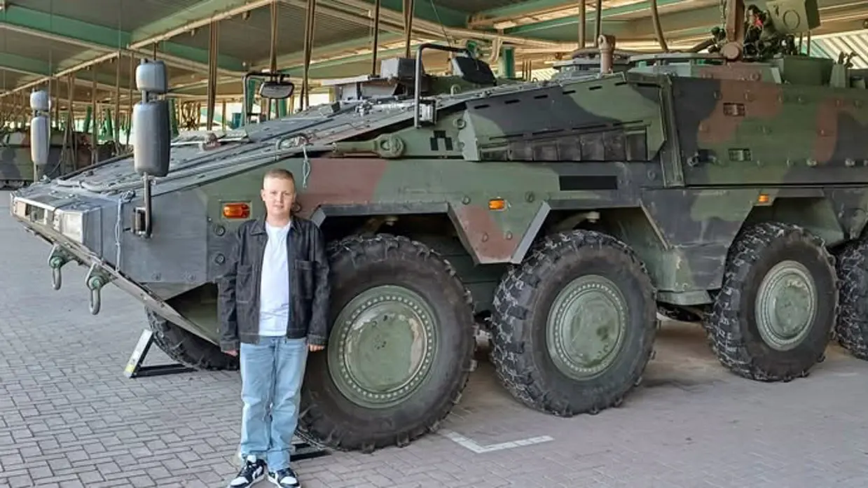 Een jongen poseert voor een groot groen pantservoertuig.