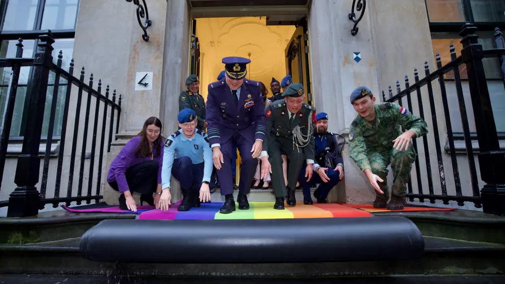 Defensiemedewerkers, onder wie de hoogste generaal, rollen een loper uit in regenboogkleuren.