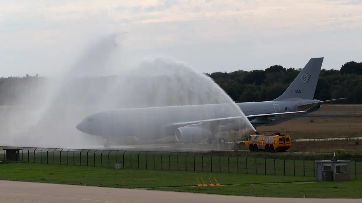 Een groot grijs vliegtuig op een startbaan wordt bespoten met water door een gele brandweerwagen; waarschijnlijk een ceremoniële waterboog.