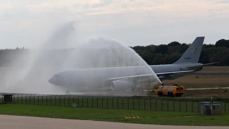 Een groot grijs vliegtuig op een startbaan wordt bespoten met water door een gele brandweerwagen; waarschijnlijk een ceremoniële waterboog.