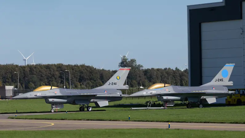 Twee grijze gevechtsvliegtuigen met gouden cockpits en blauwe staartmarkeringen staan geparkeerd op gras bij een hangar.