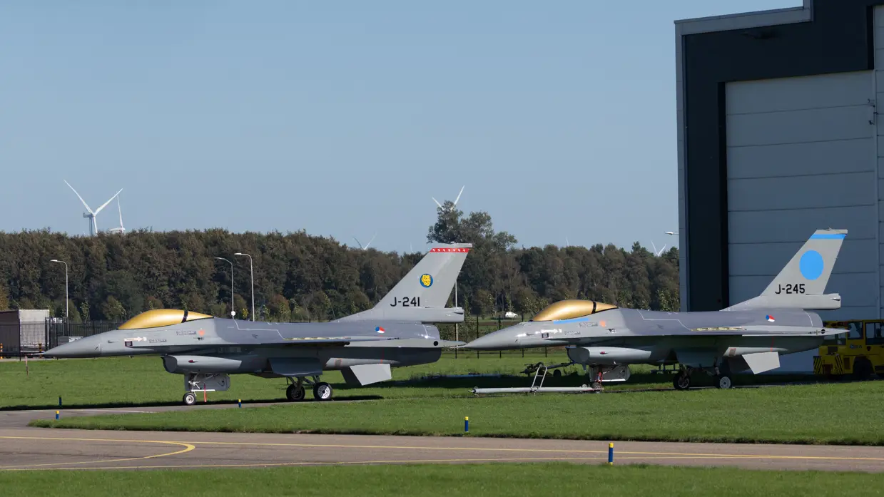 Twee grijze gevechtsvliegtuigen met gouden cockpits en blauwe staartmarkeringen staan geparkeerd op gras bij een hangar.