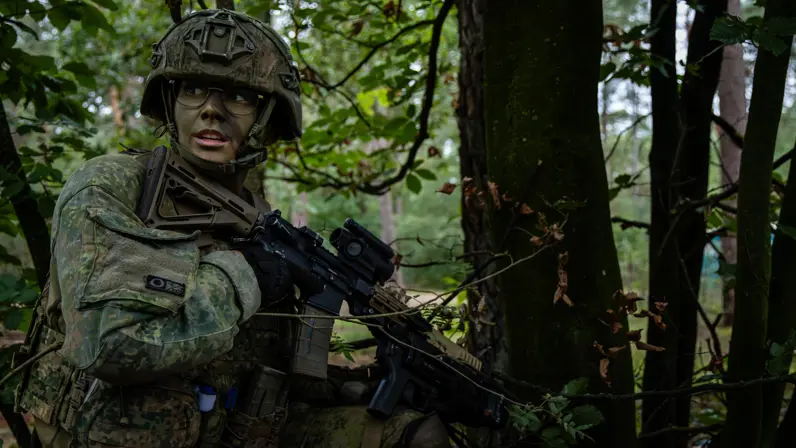 Soldaat in camouflage-uniform met helm en geweer, staand in dicht groen bos, met gefocuste, serieuze uitdrukking tijdens tactische oefening. 