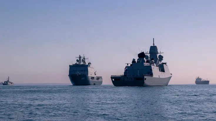 Twee Nederlandse marineschepen op een rustige zee. Op de achtergrond varen civiele transportschepen.