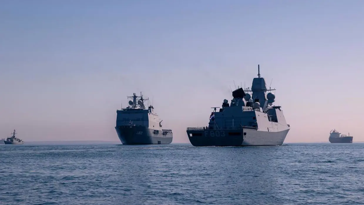Twee Nederlandse marineschepen op een rustige zee. Op de achtergrond varen civiele transportschepen.