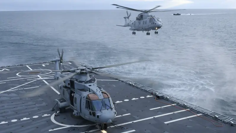Een grijze helikopter is zojuist geland op het helikopterdek van een marineschip. Daarachter hangt een tweede toestel net boven het water.