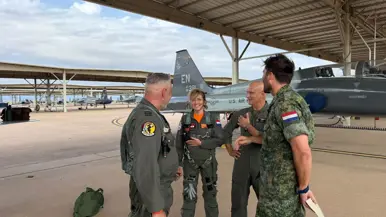De minister staat op de flightline voor een grijs vliegtuig en is in gesprek met militairen. 