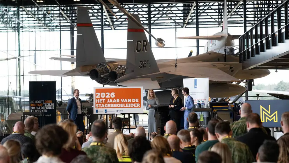 Publiek kijkt naar podium met mensen en een oranje bord. Daarachter hangt een gevechtsvliegtuig.